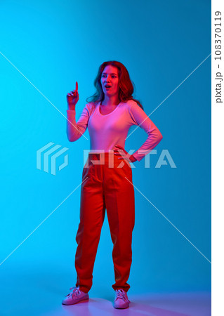 Full-length image of young girl in casual clothes raising finger up, generating ideas against blue studio background in neon light Full-length image of young girl in casual clothes raising finger up, generating ideas against blue studio background in neon light 108370119
