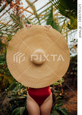 Woman in red swimsuit hides his face behind hat standing among tropical plants in greenhouse Woman in red swimsuit hides his face behind hat standing among tropical plants in greenhouse 108370125