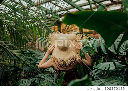 Back view of woman tourist in straw hat and leopard dress standing in palm greenhouse Back view of woman tourist in straw hat and leopard dress standing in palm greenhouse 108370136