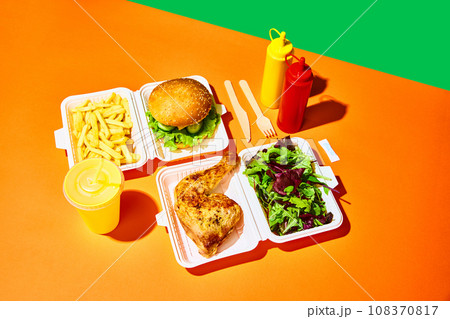 Opened paper box with salad with baked chicken, hamburger with fried potato, sauces and drinks over orange-green background. Concept of fast food, delivery. 108370817