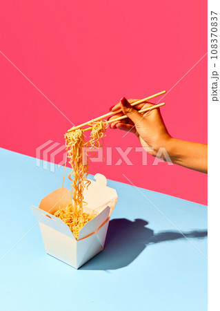 Close up image of unrecognizable girl eating from paper box with noodles, fast-food against green-pink background. Concept of fast food, delivery. Close up image of unrecognizable girl eating from paper box with noodles, fast-food against green-pink background. Concept of fast food, delivery. 108370837