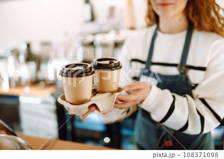 A barista woman in an apron holds hot coffee in a paper cup in her hands. 108371098