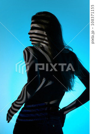 Back view of elegant young woman in jeans, looking away, posing against blue background in neon light. Stripes of shadow 108371155