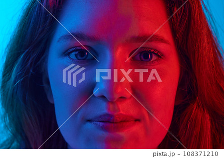 Close-up of young girl looking at camera with little smile against blue studio background in neon light Close-up of young girl looking at camera with little smile against blue studio background in neon light 108371210