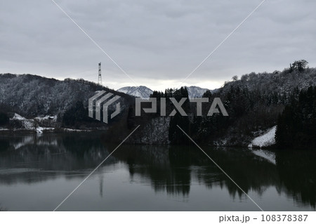 分厚い雲の奥に薄っすら明るく見える小国の雪に覆われた山々と冷たい湖畔の風景 108378387