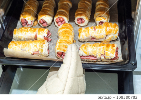 Pulling out a baking sheet with baked sausages in dough from the oven. 108380782