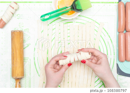 A sausage wrapped in a strip of raw dough in the hands of a young woman. 108380797