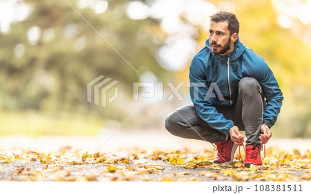 A young athlete in warm sportswear ties his shoelaces while running in an autumn park A young athlete in warm sportswear ties his shoelaces while running in an autumn park 108381511