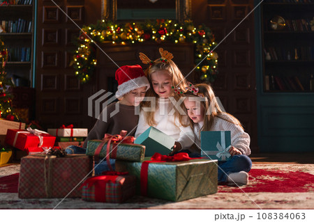 Merry Christmas Xmas holiday. Children unpacking open up gift box near Christmas tree. Kids in living room with Christmas tree fireplace opening gift box with surprise faces. Christmas eve at home 108384063