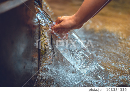 A traveler collects a bottle of cold, clean water from a spring. The man stopped to get clean water and drink from the spring. Clear, clean water flows in a stream into the offered bottle. A traveler collects a bottle of cold, clean water from a spring. The man stopped to get clean water and drink from the spring. Clear, clean water flows in a stream into the offered bottle. 108384336