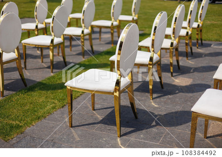 Luxury white-golden chairs on wedding ceremony outdoors. Festive decorations. Empty rows armchairs for guests. Outgoing wedding ceremony in the open air. Decor Studio. 108384492