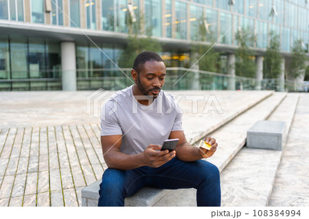 African American man shopping online holding smartphone paying with gold credit card Guy on urban street in city buying on Internet enter credit card details Online shopping ecommerce delivery service 108384994