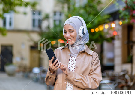 Young beautiful woman walking in the city, Muslim woman in hijab on evening walk in headphones listening to music smiling joyfully using smartphone app. Young beautiful woman walking in the city, Muslim woman in hijab on evening walk in headphones listening to music smiling joyfully using smartphone app. 108386191