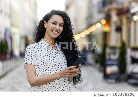 Portrait of a young beautiful Latin American woman walking in the evening city, holding a phone, smiling and looking at the camera. Portrait of a young beautiful Latin American woman walking in the evening city, holding a phone, smiling and looking at the camera. 108386200