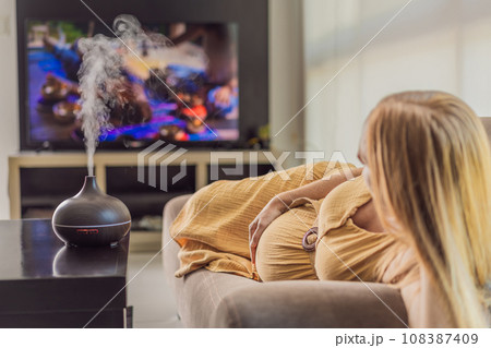 A blissful pregnant woman immerses in relaxation, savoring the soothing aroma from a diffuser while indulging in a calming TV video, embracing tranquility during her pregnant journey 108387409