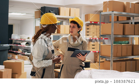 Two women with hardhats organizing products on shelves, looking at tablet to check stock logistics and planning distribution. Warehouse manager and employee counting cardboard packs. 108387621