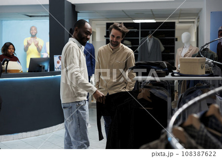 Clothing store smiling client and assistant discussing apparel style while browsing through rack. African american man showing outfit to mall customer and giving fashion advice Clothing store smiling client and assistant discussing apparel style while browsing through rack. African american man showing outfit to mall customer and giving fashion advice 108387622