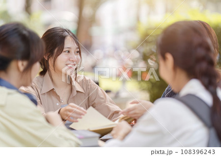 Group of young Asian college students sitting on a bench in a campus relaxation area, talking, sharing ideas, doing homework or tutoring for the exam together 108396243