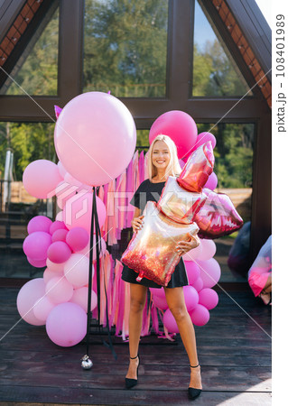 Vertical portrait of cheerful blonde female in summer dress posing holding various colorful festive foil helium balloons standing at country house building exterior, smiling looking at camera. Vertical portrait of cheerful blonde female in summer dress posing holding various colorful festive foil helium balloons standing at country house building exterior, smiling looking at camera. 108401989