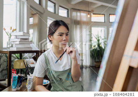 Female artist thinking and hold paintbrush while looking at canvas and pondering about paintin Female artist thinking and hold paintbrush while looking at canvas and pondering about paintin 108404281
