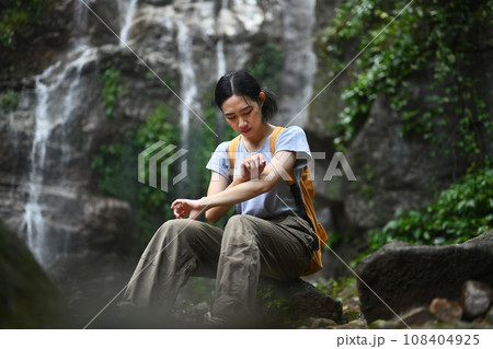 Female hiker with backpack sitting on rock in front of a waterfall resting during walking on trekking trail Female hiker with backpack sitting on rock in front of a waterfall resting during walking on trekking trail 108404925