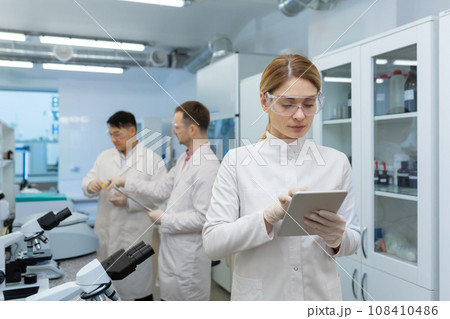 Mature confident and serious thinking woman in white medical coat working with tablet computer, team of scientists working in laboratory among microscope at workplace inside. 108410486