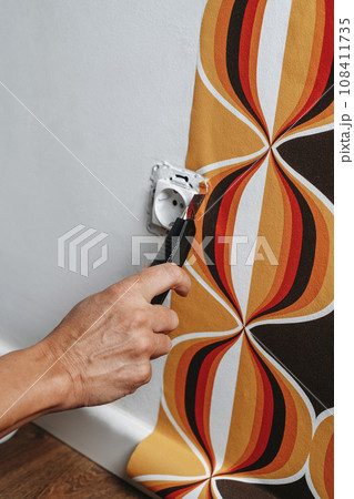 man cuts the edges of a wallpaper around a socket man cuts the edges of a wallpaper around a socket 108411735