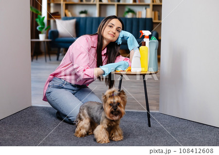 Young woman leaning on the table near cleaning products at home and her little dog is standing next to her Young woman leaning on the table near cleaning products at home and her little dog is standing next to her 108412608