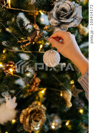 Close-up vertical cropped shot of unrecognizable happy little girl putting xmas toys on illuminated xmas tree branches. Closeup of kid decorating illuminated Christmas tree with balls. Close-up vertical cropped shot of unrecognizable happy little girl putting xmas toys on illuminated xmas tree branches. Closeup of kid decorating illuminated Christmas tree with balls. 108414172
