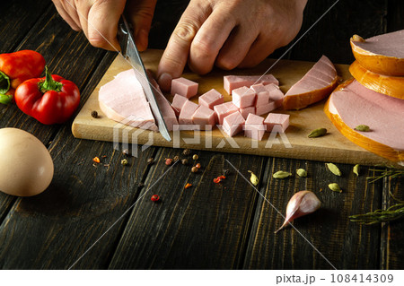 Slicing parboiled sausage with a knife in the hands of a chef to prepare a delicious salad. Concept of preparing food for the holiday table 108414309