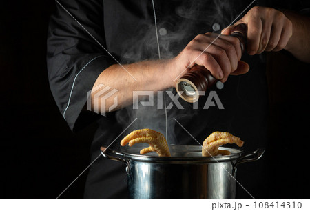 The cook adds dry pepper to a pot of boiling chicken legs. Concept of preparing delicious soup or broth in the kitchen of a restaurant or hotel 108414310