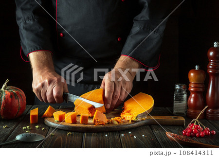 A man cuts a pumpkin with a kitchen knife on a wooden board. Fresh raw diced pumpkin on a kitchen cutting board. Preparing ingredients for a seasonal fall dish at home 108414311