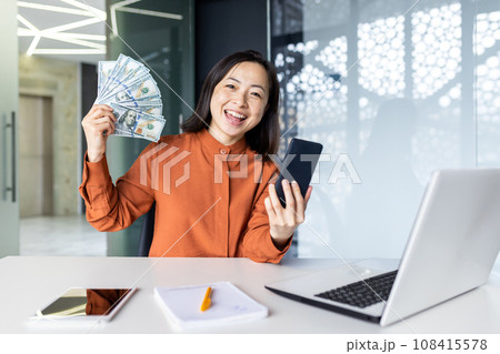 Portrait of young joyful asian business woman, woman smiling and looking at camera, holding phone and winning money cash american dollars, working at workplace inside office. Portrait of young joyful asian business woman, woman smiling and looking at camera, holding phone and winning money cash american dollars, working at workplace inside office. 108415578