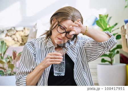 Mature woman suffering from headache, at home with glass of water Mature woman suffering from headache, at home with glass of water 108417951