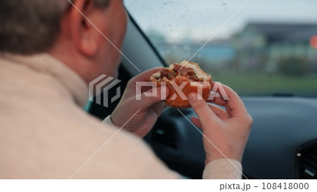 Man takes bite of tasty burger to quell hunger sitting in car on cloudy day. Man consuming burger in car and enjoying view from window. Man indulging in unhealthy burger in car in move on road of city 108418000