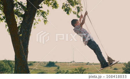 Boy enthusiastically swings on homemade swing enjoying time in nature. Boy swings energetically on swing spending time in natural setting. Boy spends time in nature swinging back and forth on swing Boy enthusiastically swings on homemade swing enjoying time in nature. Boy swings energetically on swing spending time in natural setting. Boy spends time in nature swinging back and forth on swing 108418033