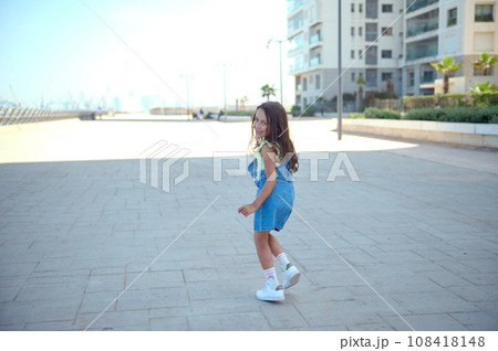Little girl in casual denim dress, smiling looking at the camera through her shoulders, running along marine promenade 108418148