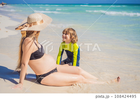A radiant pregnant mother and her excited son share a tender moment on a serene, snow-white beach, celebrating family love amidst nature's beauty 108424495