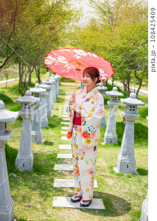 Young woman wearing a Japanese traditional kimono or yukata holding an umbrella in a garden with torii poles. 108425409