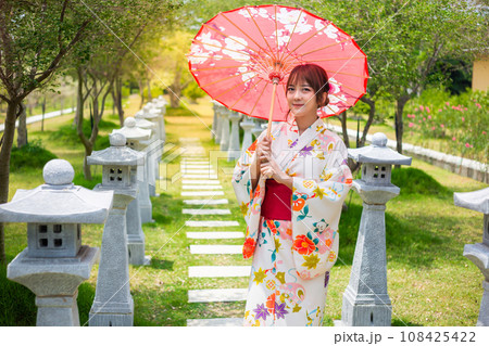 Young woman wearing a Japanese traditional kimono or yukata holding an umbrella in a garden with torii poles. Young woman wearing a Japanese traditional kimono or yukata holding an umbrella in a garden with torii poles. 108425422