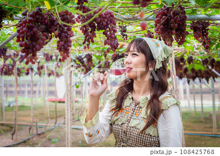 Woman wearing cute dress stand drinking grape juice or wine in vineyard smiling happily Woman wearing cute dress stand drinking grape juice or wine in vineyard smiling happily 108425768