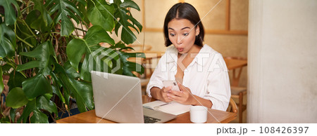 Woman in cafe sitting with laptop, looking surprised at smartphone screen, reading exciting good news 108426397