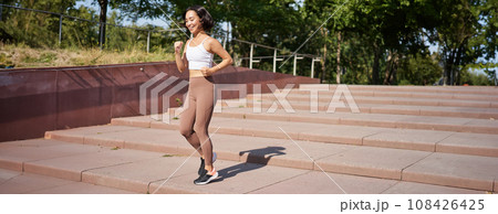 Portrait of young sportswoman running on street. Fitness girl jogging in park, workout outdoors, listening music in wireless earphones 108426425