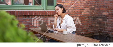 Beautiful brunette woman sitting outdoors and using laptop, chatting online, working with computer outside office 108426507