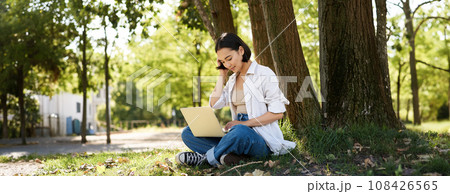 Remote work. Smiling asian girl, student doing homework remotely from park, sitting with laptop near tree and typing 108426565
