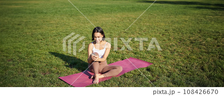 Woman in park, watching yoga video on smartphone, meditating on fresh air, sitting on rubber mat Woman in park, watching yoga video on smartphone, meditating on fresh air, sitting on rubber mat 108426670