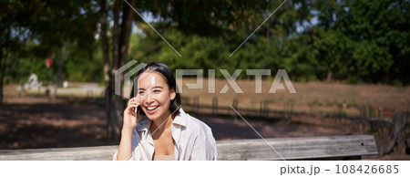 Portrait of happy young asian woman talking on mobile phone in park, sitting on bench and having a telephone call, chatting lively Portrait of happy young asian woman talking on mobile phone in park, sitting on bench and having a telephone call, chatting lively 108426685