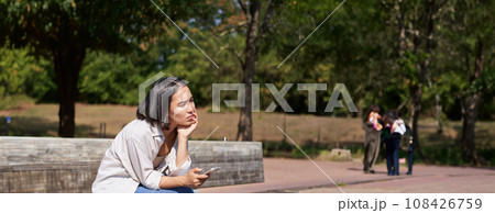 Portrait of asian girl sitting with smartphone feeling sad, looking gloomy and frustrated, waiting for a call outdoors in park Portrait of asian girl sitting with smartphone feeling sad, looking gloomy and frustrated, waiting for a call outdoors in park 108426759