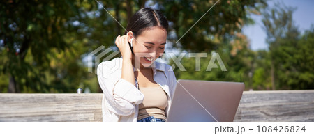 Beautiful asian girl laughing, watching video on laptop, listening music or video chatting online while sitting in park on bench 108426824