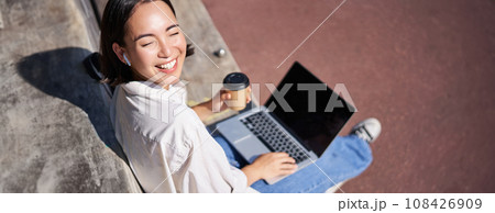 Beautiful young asian female student, sitting with laptop on bench in park, drinking takeaway coffee and listening music, working remotely, studying outdoors 108426909